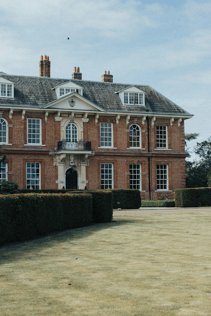 Photo of a large mansion in the UK countryside, with a hedge and large area of grass in front of the front door.